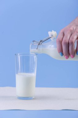 Bir kadın eli mavi bir arka plan üzerine bir bardak içine bir cam şişe taze süt dökülen. Renkli minimalizm. Kalsiyum sağlıklı süt ürünleri kavramı
