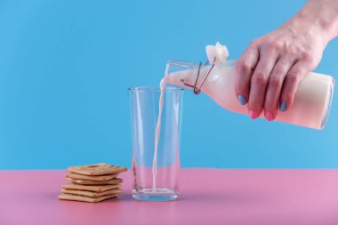 Bir kadın eli bir cam ve çerezleri pastel bir arka plan üzerine içine bir cam şişe taze süt dökülen. Renkli minimalizm. Kalsiyum sağlıklı süt ürünleri kavramı