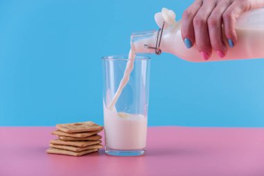Bir kadın eli bir cam ve çerezleri pastel bir arka plan üzerine içine bir cam şişe taze süt dökülen. Renkli minimalizm. Kalsiyum sağlıklı süt ürünleri kavramı