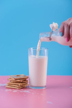 Bir kadın eli bir cam ve çerezleri pastel bir arka plan üzerine içine bir cam şişe taze süt dökülen. Renkli minimalizm. Kalsiyum sağlıklı süt ürünleri kavramı