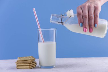 Bir kadın eli bir cam ve çerezleri mavi bir arka plan üzerinde bir cam şişe taze süt dökülen. Renkli minimalizm. Kalsiyum sağlıklı süt ürünleri kavramı