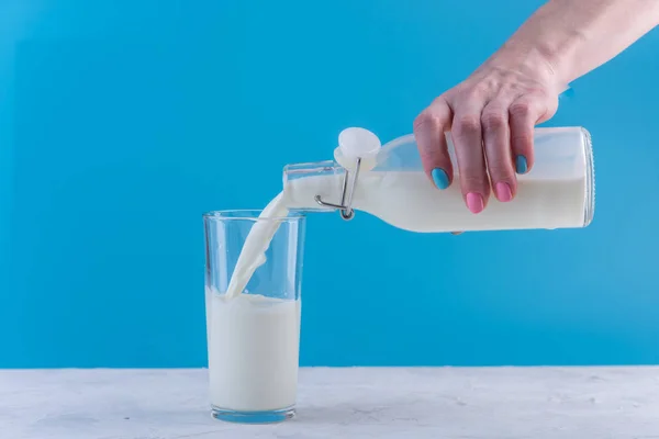 A woman\'s hand pours fresh milk from a glass bottle into a glass on a blue background. Colorful minimalism. The concept of healthy dairy products with calcium