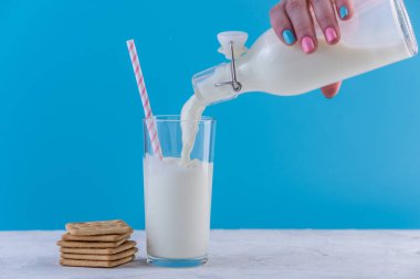 Bir kadın eli bir cam ve çerezleri mavi bir arka plan üzerinde bir cam şişe taze süt dökülen. Renkli minimalizm. Kalsiyum sağlıklı süt ürünleri kavramı