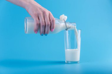 Bir kadın eli mavi bir arka plan üzerine bir bardak içine bir cam şişe taze süt dökülen. Renkli minimalizm. Kalsiyum sağlıklı süt ürünleri kavramı