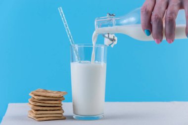 Bir kadın eli bir cam ve çerezleri mavi bir arka plan üzerinde bir cam şişe taze süt dökülen. Renkli minimalizm. Kalsiyum sağlıklı süt ürünleri kavramı