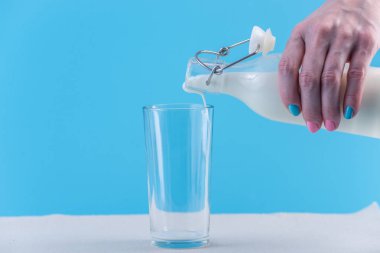 Bir kadın eli mavi bir arka plan üzerine bir bardak içine bir cam şişe taze süt dökülen. Renkli minimalizm. Kalsiyum sağlıklı süt ürünleri kavramı