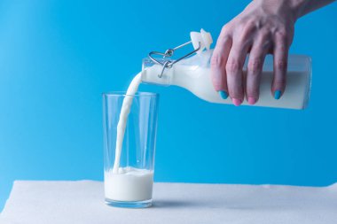 Bir kadın eli mavi bir arka plan üzerine bir bardak içine bir cam şişe taze süt dökülen. Renkli minimalizm. Kalsiyum sağlıklı süt ürünleri kavramı