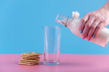 Bir kadın eli bir cam ve çerezleri pastel bir arka plan üzerine içine bir cam şişe taze süt dökülen. Renkli minimalizm. Kalsiyum sağlıklı süt ürünleri kavramı