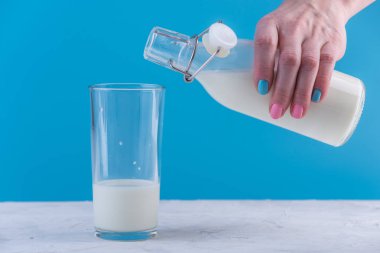 Bir kadın eli mavi bir arka plan üzerine bir bardak içine bir cam şişe taze süt dökülen. Renkli minimalizm. Kalsiyum sağlıklı süt ürünleri kavramı
