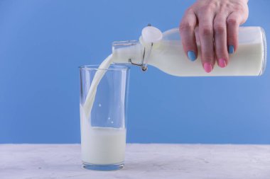Bir kadın eli mavi bir arka plan üzerine bir bardak içine bir cam şişe taze süt dökülen. Renkli minimalizm. Kalsiyum sağlıklı süt ürünleri kavramı