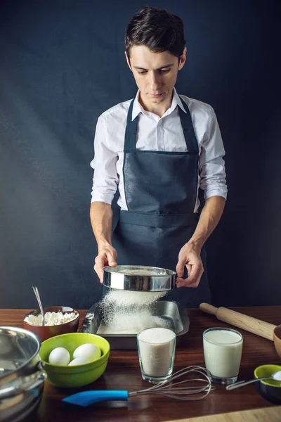 Young man chef in uniform black apron cooking cake with ingredients on the table in the kitchen. The concept of men cook desserts