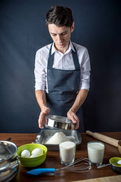 Young man chef in uniform black apron cooking cake with ingredients on the table in the kitchen. The concept of men cook desserts