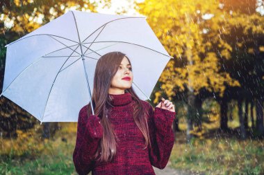 Mutlu genç kadın kırmızı kazaklı bir Sunny Park'ta yağmur altında beyaz bir şemsiye ile yürür. Mevsim ve sonbahar ruh kavramı