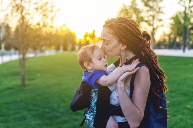 Genç mutlu anne bebek oğlu ile ergo sırt çantası Park'ta yürüyordunuz. Güneşli yaz günü. Modern anne kavramı