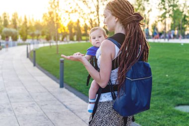Genç mutlu anne bebek oğlu ile ergo sırt çantası Park'ta yürüyordunuz. Güneşli yaz günü. Modern anne kavramı