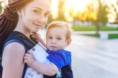 Genç mutlu anne bebek oğlu ile ergo birlikte seyahat etmek sırt çantası. Güneşli yaz günü parkta. Modern anne kavramı