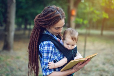 Mutlu anne parkta bir kitap okuma bir askı içinde bir bebekle. Aile hayatında çocuk eğitim kavramı