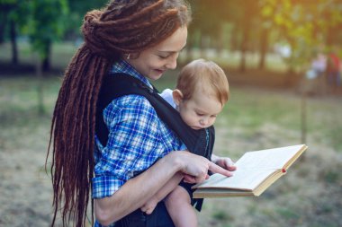 Mutlu anne parkta bir kitap okuma bir askı içinde bir bebekle. Aile hayatında çocuk eğitim kavramı