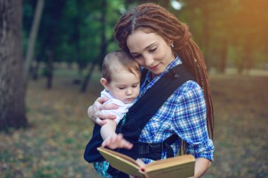 Mutlu anne parkta bir kitap okuma bir askı içinde bir bebekle. Aile hayatında çocuk eğitim kavramı