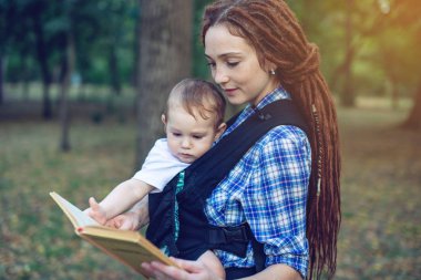 Mutlu anne parkta bir kitap okuma bir askı içinde bir bebekle. Aile hayatında çocuk eğitim kavramı