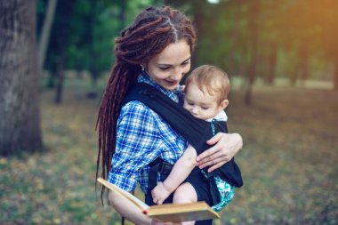 Bir bebek ile güzel anne ergo sırt çantası parkta bir masal okuma. Çocuk eğitiminde aile hayatı
