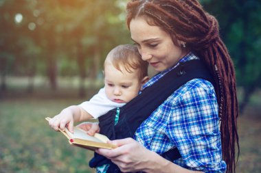 Bir bebek ile güzel anne ergo sırt çantası parkta bir masal okuma. Çocuk eğitiminde aile hayatı