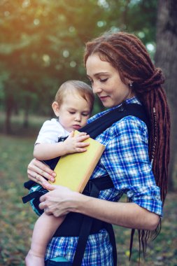 Bir bebek ile güzel anne ergo sırt çantası parkta bir masal okuma. Çocuk eğitiminde aile hayatı