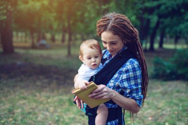 Bir bebek ile güzel anne ergo sırt çantası parkta bir masal okuma. Çocuk eğitiminde aile hayatı