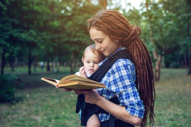 Bir bebek ile güzel anne ergo sırt çantası parkta bir masal okuma. Çocuk eğitiminde aile hayatı