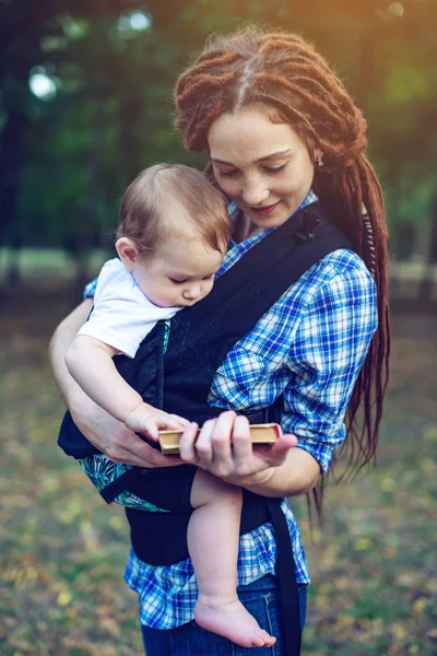Bir bebek ile güzel anne ergo sırt çantası parkta bir masal okuma. Çocuk eğitiminde aile hayatı