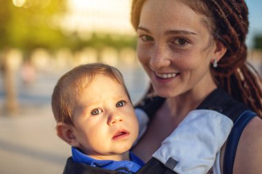 Genç mutlu anne bebek oğlu ile ergo birlikte seyahat etmek sırt çantası. Arka planı karanlık duvara. Modern anne kavramı