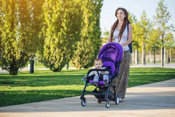 Young modern mom with baby son in stroller walking in Sunny Park. The concept of the joy of motherhood and autumn mood