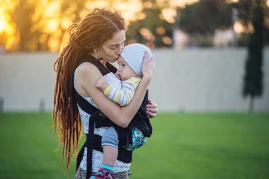 Genç modern mutlu anne bebek oğlu ile ergo sırt çantası içinde güneşli yaz günü yürüyüş. Annelik sevinci kavramı