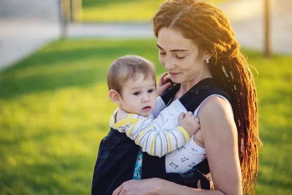 Genç modern mutlu anne bebek oğlu ile ergo sırt çantası içinde güneşli yaz günü yürüyüş. Annelik sevinci kavramı