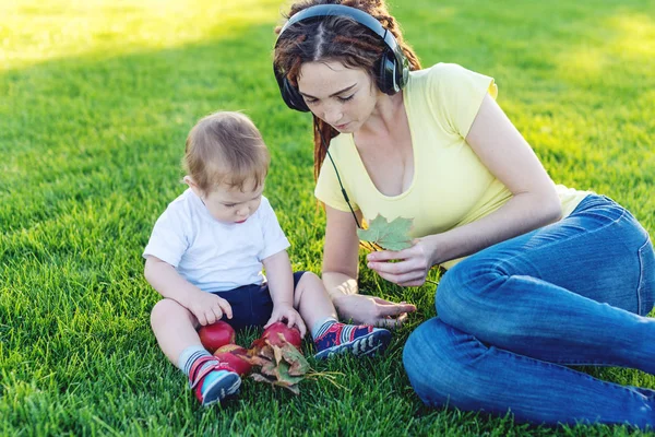 Beautiful young modern mom in headphones playing in a green meadow with her cute baby son in a Sunny Park. The concept of the joy of motherhood