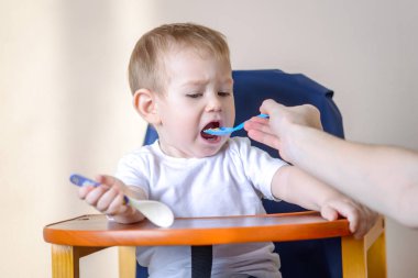 Küçük oğlu ağzını mutfakta bir sandalyede oturan geniş açılış yiyor. Anne elinde tutan bir kaşık yulaf lapası beslemeleri