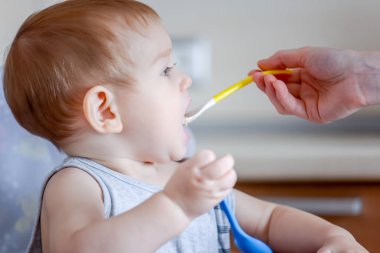 Küçük oğlu ağzını mutfakta bir sandalyede oturan geniş açılış yiyor. Anne elinde tutan bir kaşık yulaf lapası beslemeleri