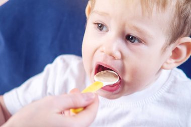 Küçük şirin bebek çocuk ağzını mutfakta bir sandalyede oturan geniş açılış yeme. Anne elinde tutan bir kaşık yulaf lapası beslemeleri