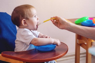 Küçük şirin bebek çocuk ağzını mutfakta bir sandalyede oturan geniş açılış yeme. Anne elinde tutan bir kaşık yulaf lapası beslemeleri