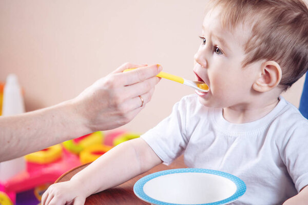 Little cute baby boy eating on a chair in the kitchen. Mom feeds holding in hand a spoon of porridge