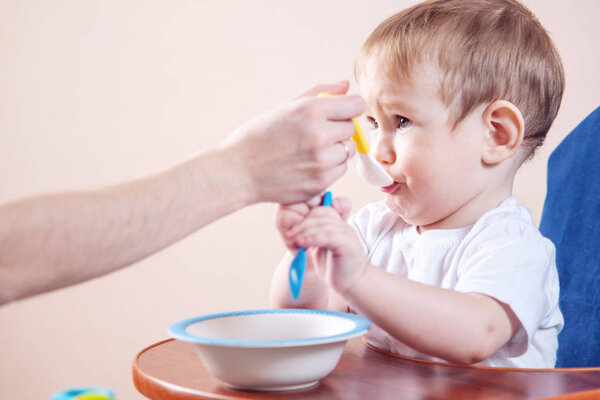 Little cute baby boy eating on a chair in the kitchen. Mom feeds holding in hand a spoon of porridge