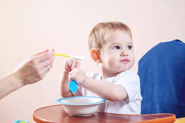 Little cute baby boy eating on a chair in the kitchen. Mom feeds holding in hand a spoon of porridge