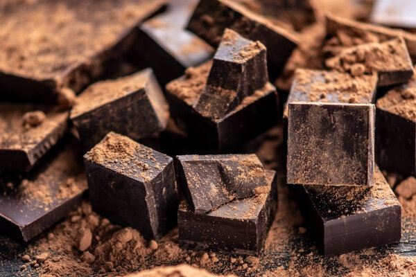 Кусочки тёмного горького шоколада с какао-порошком на тёмном фоне. Концепция кондитерских изделий

