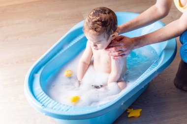 Anne küçük çocuk banyoda mavi bir banyoda yıkama. Sarı ördek oyuncak ve sabun köpüğü ile oynayan bebek.