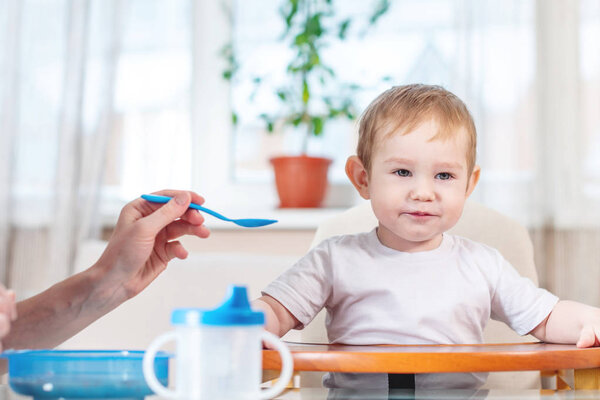Mother feeding the baby holding out her hand with a spoon of porridge in the kitchen. Emotions of a child while eating healthy food.