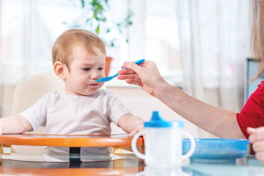 Mother feeding the baby holding out her hand with a spoon of porridge in the kitchen. Emotions of a child while eating healthy food.