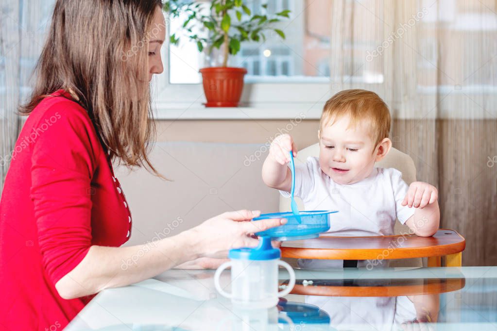 Mom feeding the baby holding out her hand with a spoon of food in the kitchen. Healthy baby nutrition. The emotions of a child while eating