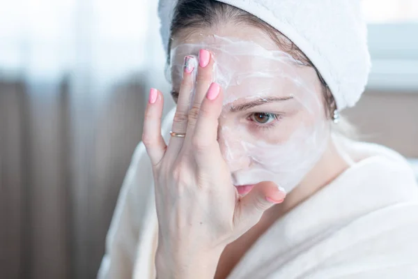 Beautiful happy young woman with a towel on her head applying cream of natural cosmetics on face moisturizing the skin. The concept of hygiene and care for the skin at home