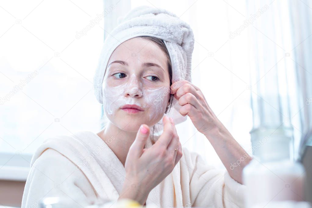 Beautiful happy young woman with a towel on her head applying cream of natural cosmetics on face moisturizing the skin. The concept of hygiene and care for the skin at home