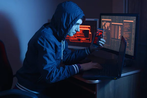 Hacker in the hood holding the phone in his hands trying to hack the mobile device cloud and steal data in the dark under neon light. The concept of cyber security